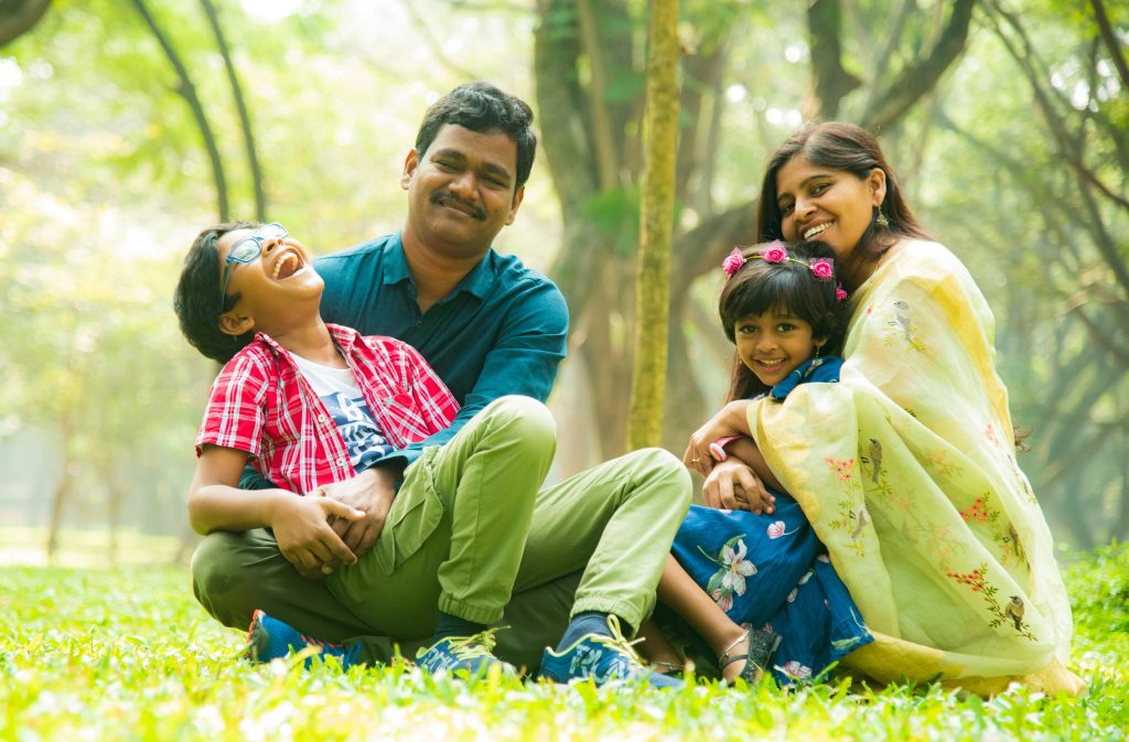 A happy mother, father, and their two children sitting closely on the ground, smiling and hugging to represent family wellbeing and emotional connection.